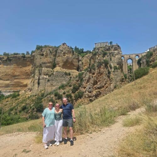 Tourists standing on a trail with cliffs and ancient ruins in the background.