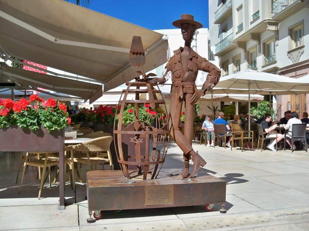 Statue of a traditional Málaga figure holding a wine barrel, located outdoors at a lively café area.